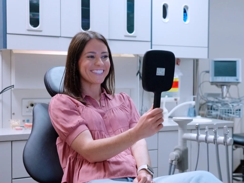 Young woman smiling in mirror after straightening her teeth with clear aligner treatment at Conroe Dental Associates in Conroe, TX
